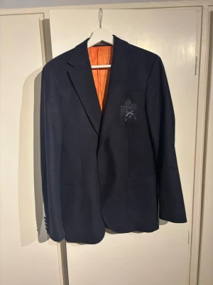 Mörkblå kavaj med från Morris - Tjena! Säljer en mörkblå kavaj från Morris med orange innerfoder och ett broderat emblem på bröstfickan. Skick 9/10. Storlek 44. Bara att fråga om det är något!