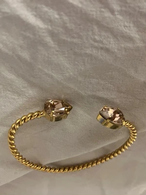 Guldarmband från Caroline svedblom - Snyggt guldarmband från Caroline  svedblom med tvinnad design och två stora, ljusa kristaller i ändarna. Armbandet är öppet och har en exklusiv känsla, perfekt för att lyfta vilken outfit som helst.