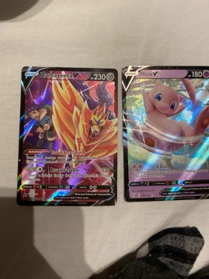 Pokémonkort Zamazenta & Mew V - Två Pokémonkort: Zamazenta V och Mew V. Båda korten har holografisk yta med färgstarka illustrationer. Zamazenta-kortet har en röd och gul design med en sköld, medan Mew-kortet är lila och blått med en söt Mew. Perfekt för samlare eller Pokémonfans.