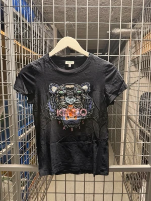 Svart Kenzo t-shirt med tigertryck - Cool svart t-shirt från Kenzo med ikoniskt tigertryck och färgglada detaljer framtill. Klassisk rund halsringning och korta ärmar. Perfekt statement-plagg för dig som vill sticka ut.