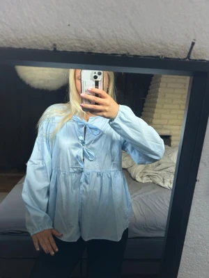 Ljusblå blus med rosettknäppning - Söt ljusblå blus med två rosetter framtill och avslappnad passform. Blusen har långa ärmar med resår vid handlederna och ett lätt glansigt tyg som ger en fräsch look. 