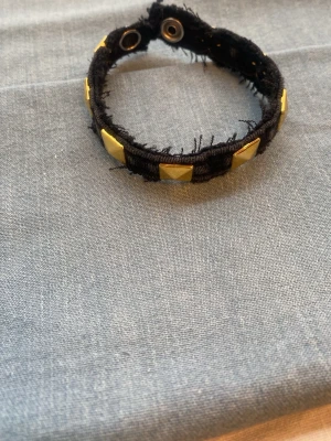 Svart tygarmband med nitar - Coolt svart armband i tyg med råa kanter och guldiga pyramidformade nitar. Stängs med tryckknappar och har en edgy, trendig look som passar perfekt till streetstyle eller rockiga outfits.