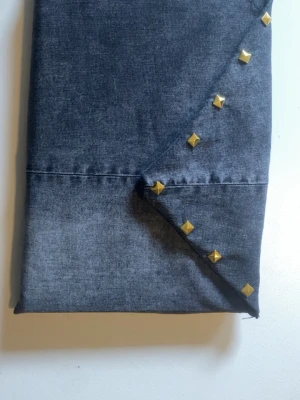 Laptopfodral i denim med nitar - Snyggt laptopfodral i mörkgrå denim med guldfärgade fyrkantiga nitar längs kanten. Fodralet har en enkel, vikbar design och är gjort av tygmaterial som ger en cool och stilren look. Perfekt för att skydda din dator på språng.