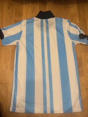 Argentina fotbollströja Adidas - Säljer en klassisk Argentina fotbollströja från Adidas med blå och vita vertikala ränder, svart krage och AFA-emblem på bröstet. Tröjan är kortärmad och tillverkad i lätt och ventilerande material med Aeroready-teknologi.