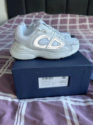 Vita Dior B30 sneakers med logga - Snygga vita Dior B30 sneakers med stor CD-logga på sidan och bak. Skorna har en chunky sula och är tillverkade i mesh och läder för en modern look. Perfekta för dig som vill ha exklusiva och trendiga sneakers med premiumkänsla.