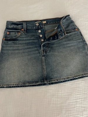 Blå jeanskjol levis  - Snygg blå jeanskjol från Icon Skirt med klassisk femficksdesign och knappar framtill. Kjolen har en kort passform och är tillverkad i slitstarkt denimtyg. Perfekt för dig som gillar en tidlös och avslappnad stil.