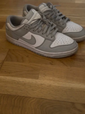 Nike Dunk Low grå och vita sneakers - Snygga Nike Dunk Low sneakers i vitt och ljusgrått läder med klassisk swoosh på sidorna. Låg modell med perforerad tåbox och platt sula. Perfekta för dig som gillar stilrena och tidlösa sneakers med streetkänsla.