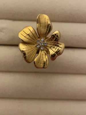 Guldfärgad blomring med stenar - Unik ring i form av en blomma med fem blad i guldfärgad metall. I mitten sitter små gnistrande stenar som ger extra bling. Perfekt för dig som vill sticka ut med ett coolt och trendigt smycke. Blommans detaljerade blad ger ringen en lyxig känsla. Rostfri 