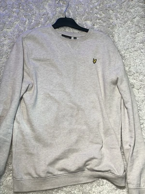Ljusgrå sweatshirt från Lyle & Scott lite beige  - En stilren ljusgrå sweatshirt från Lyle & Scott med klassisk rund hals och det ikoniska gula örnloggan broderad på bröstet. Tröjan har långa ärmar och är gjord i mjukt bomullsmaterial, perfekt för en avslappnad och trendig look.