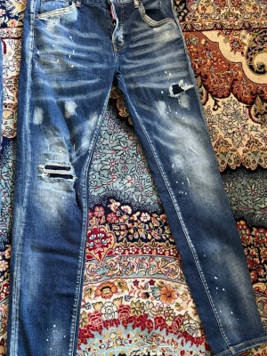 Blå slitna jeans från Dsquared2 - Säljer ett par blå jeans från Dsquared2 med slitningar, hål och färgstänk för en riktigt edgy look. Jeansen har klassisk femficksmodell, knappgylf och smal passform. Tillverkade i denim med cool tvätt och detaljer som sticker ut.