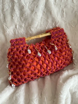 orange handväska med pärlor - Unik handväska i orange tyg med virkat nät i lila och orange. Väskan har handtag och dekoreras med hängande vita pärlor. Perfekt statement piece för dig som gillar färg och detaljer. Väskan har en rektangulär form och är handgjord.