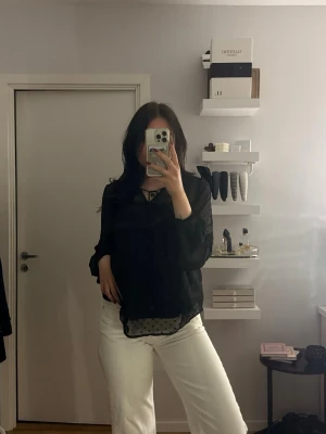 Svart blus med spetsdetaljer - Svart blus med långa ärmar och fina spetsdetaljer. Blusen har en lös passform och knytning vid halsen. Materialet ser lätt och något transparent ut, perfekt för en trendig look.