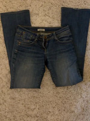Blå bootcut jeans - Säljer ett par klassiska blå bootcut jeans  med snygga slitningar och femficksmodell. Jeansen har låg midja och gylf med dragkedja och knapp.           Xs-s