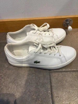 Vita Lacoste sneakers i läder - Klassiska vita sneakers från Lacoste i slät läder med snörning och den ikoniska gröna krokodilloggan på sidan. Snygg och stilren design med lågt skaft och vit sula. Perfekta för en clean och sportig look.