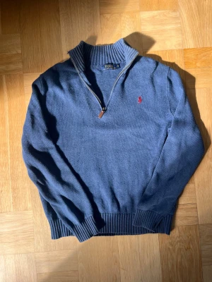 Blå stickad tröja från Polo Ralph Lauren - Mysig blå stickad tröja från Polo Ralph Lauren med halv dragkedja och hög krage. Tröjan har ribbade muddar och den klassiska röda broderade loggan på bröstet. Perfekt för lager-på-lager och chill stil.