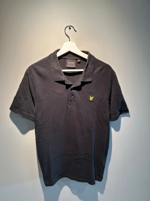 Svart pikétröja från Lyle & Scott - Klassisk svart pikétröja från Lyle & Scott med gul broderad logga på bröstet. Tröjan har krage, knappar framtill och korta ärmar. Perfekt för en stilren och avslappnad look. Storlek L.