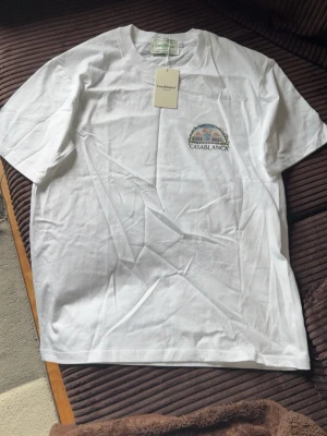 Vit t-shirt från Casablanca - Vit t-shirt från Casablanca med tryck av en byggnad och logga på bröstet. Klassisk rund hals och korta ärmar. Tillverkad i mjuk bomull som känns skön mot huden. Perfekt för en clean och stilren look.