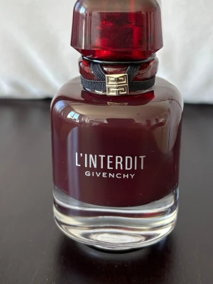 Givenchy L'Interdit parfym mörkröd - Givenchy L'Interdit parfym i en elegant mörkröd glasflaska med genomskinlig botten och matchande röd kork. Flaskan har en svart dekorativ banddetalj med metallisk logga runt halsen. Perfekt för dig som gillar lyxiga och stilrena dofter.
