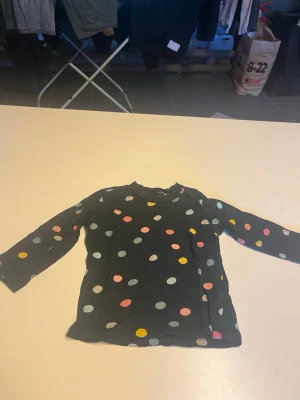 Svart långärmad topp med prickar - Svart långärmad topp med färgglada prickar i rosa, blått, gult och vitt. Mjuk och skön passform, perfekt för dig som gillar lekfulla mönster. Rund halsringning och stretchigt material.