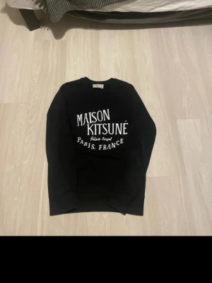 Maison Kitsuné svart hoodie - Svart hoodie från Maison Kitsuné med vit tryckt logga och text på bröstet. Klassisk passform med rund hals och ribbade muddar. Perfekt för dig som gillar stilrena och trendiga plagg med fransk vibe.