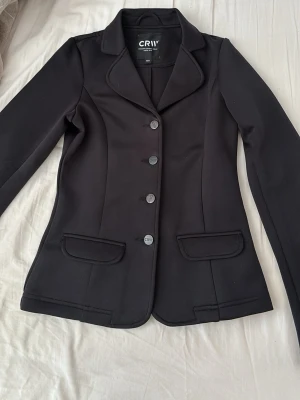 Svart kavaj från CRW med fickor - Stilren svart kavaj från CRW med klassisk krage, fyra knappar framtill och två fickor med lock. Figursydd modell som ger en snygg siluett. Perfekt för dig som vill ha en snygg outfit på en rid tävling!
