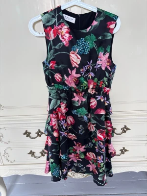 Blommig kortklänning från By Malina - Snygg ärmlös kortklänning (1 dm ovanför knät) från By Malina med färgstarkt blommönster på svart botten. Klänningen har markerad midja med resår och ett mjukt, glansigt material som ger en lyxig känsla. Perfekt för dig som vill sticka ut med färg och mönster. Klänning är aldrig använd utan hängt i garderoben efter att taggen klipptes bort.
