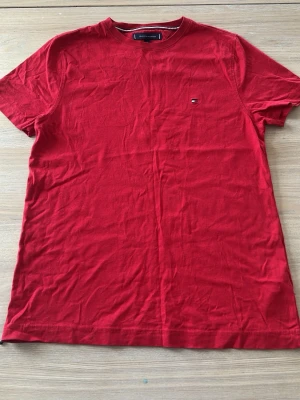 Röd t-shirt från Tommy Hilfiger - Snygg röd t-shirt från Tommy Hilfiger med klassisk passform och liten broderad logga på bröstet. T-shirten har rund halsringning och är tillverkad i mjuk bomull, perfekt för en avslappnad stil.