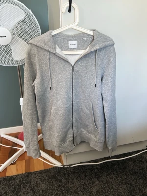 Grå hoodie med dragkedja Jack & Jones - Snygg grå hoodie från Jack & Jones med dragkedja framtill, huva med dragsko och två stora fickor. Tillverkad i mjukt material som känns skönt mot huden. Perfekt för en avslappnad stil.