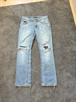 Sällsynta Nudie jeans - Ljusblå jeans med raka ben, slitningar och hål vid knäna samt fransade benslut. Klassisk femficksmodell med låg midja och detaljerade bakfickor. Perfekta för en avslappnad och trendig streetstil. Modell: Bootcut Ola (discontinued fit)