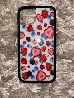Wildflower case iPhone 15 plus - Färgglatt mobilskal med jordgubbar, blåbär och björnbär på vit bakgrund. Skalet har en svart ram och är gjort i hårdplast. Perfekt för dig som vill ha ett unikt och lekfullt skydd till mobilen.