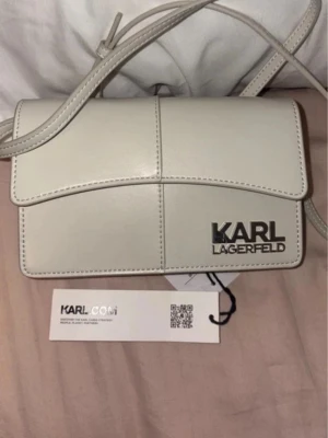 Vit axelremsväska från Karl Lagerfeld - Snygg vit handväska från Karl Lagerfeld i slätt konstläder. Väskan har en stilren, rektangulär form med silverfärgad logotyp framtill och justerbar axelrem. Perfekt för dig som gillar minimalistisk och trendig design.