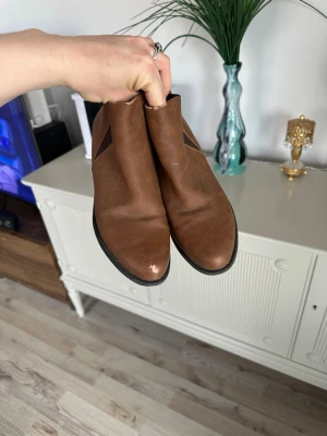 Bruna klassiska stövletter i skinnimitation - Se bilder för skick!! Snygga bruna stövletter med låg klack och elastiska partier på sidorna för enkel påtagning. Skorna har en stilren design med rundad tå och svart sula. Perfekta för dig som gillar en enkel och tidlös look.