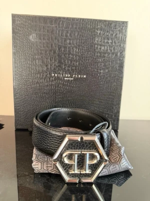 Svart skärp från Philipp Plein - Snyggt svart skärp från Philipp Plein med en stor, sexkantig metallspänne med PP-logga. Skärpet är gjort i präglat skinn och har en exklusiv känsla. Kommer med originalbox och dustbag. 100cm långt. Nypris 4500kr