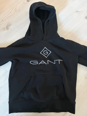 Svart hoodie från GANT med logga - Svart hoodie från GANT med stor vit broderad logga på bröstet och klassisk känguruficka. Mjuk insida och huva för extra komfort. Perfekt för en avslappnad och stilren look.