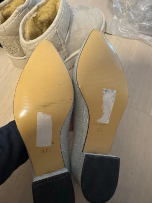 Silvriga glittriga pumps med låg klack - Säljer ett par silvriga pumps med glittrigt tyg och låg, bred klack. Skorna har spetsig tå och en klassisk, elegant form som passar perfekt till festliga tillfällen. Innersulan är beige och yttermaterialet skimrar snyggt i ljuset.