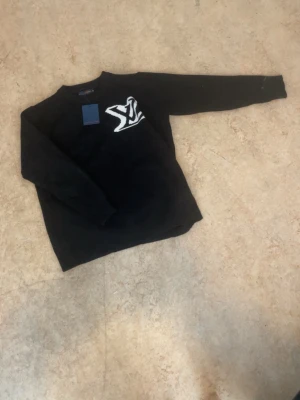 Svart tröja med vit logga - Svart långärmad tröja med en stor vit broderad logga på bröstet. Tröjan har rund halsringning och är tillverkad i ett mjukt material som känns skönt mot huden. Perfekt för en avslappnad och stilren look.