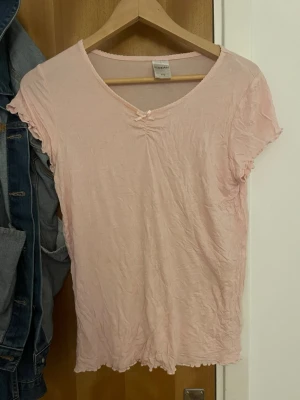 Ljusrosa t-shirt med rosett - Supersöt ljusrosa t-shirt från kapahlmed liten rosett vid halsringningen och volangkant på ärmar och nederkant. T-shirten har korta ärmar och är gjord i mjuk bomull, perfekt för en chill och avslappnad look.