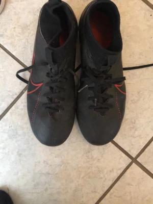 Svarta Nike fotbollsskor med röd detalj - Säljer ett par svarta fotbollsskor från Nike med röda detaljer och klassisk Nike-logga på sidorna. Skorna har snörning och dobbar för bra grepp på planen. Snygg och sportig design som passar perfekt för matcher och träning.