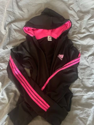 Svart Adidas hoodie med rosa detaljer - Svart hoodie från Adidas med dragkedja, rosa foder i huvan och rosa ränder längs ärmarna. Adidas-logga i rosa på bröstet. Sportig och snygg design, perfekt för en avslappnad stil. Tillverkad i ett mjukt och stretchigt material.