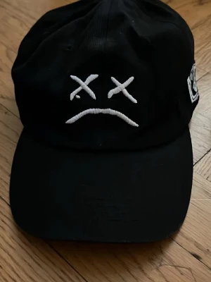 Svart keps med broderad smiley - Säljer en svart keps med böjd skärm och broderad vit smiley med korsade ögon och nedåtvänd mun på framsidan. Kepsen har en patch på sidan och är tillverkad i bomull. Perfekt för en streetwear-look.