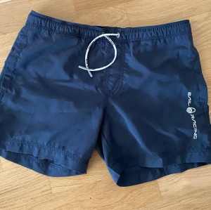Marinblå badbyxor från Sail Racing - Snygga marinblå badbyxor från Sail Racing med vit dragsko i midjan och logotyp tryckt på ena benet. Klassisk modell med kortare ben och elastisk midja. Perfekta för stranden eller poolen.