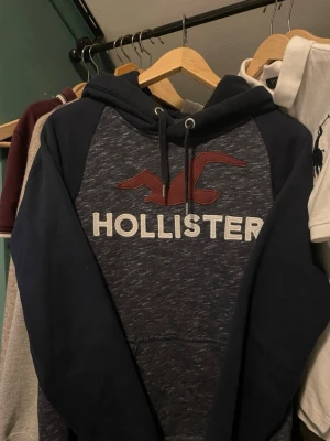 Mörkblå Hollister hoodie med logga - Snygg mörkblå hoodie från Hollister med stor vit logga och röd fågel på bröstet. Tröjan har huva med dragsko och är tillverkad i mjukt, fleeceliknande material. Perfekt för en avslappnad och trendig stil.
