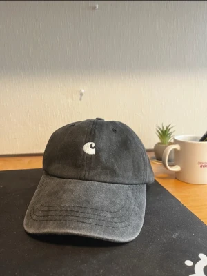 Svart keps från Carhartt - Snygg svart keps från Carhartt med böjd skärm och broderad vit logga framtill. Klassisk dad cap-modell i bomullstyg, perfekt för en avslappnad streetwear-look.