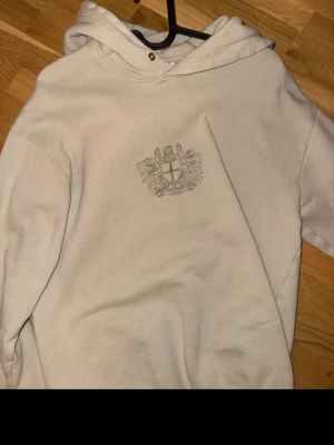Beige hoodie från Han Kjøbenhavn - Snygg beige hoodie från Han Kjøbenhavn med broderat emblem på bröstet. Hoodien har en klassisk passform, huva med metallöljetter och är tillverkad i mjukt material som känns skönt mot huden.