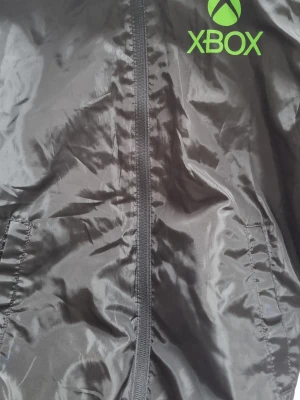 Svart vindjacka med Xbox-logga - Snygg svart vindjacka med dragkedja framtill och två sidofickor. Jackan har en grön Xbox-logga tryckt på bröstet och är tillverkad i ett glansigt, lätt material som passar perfekt för blåsiga dagar.
