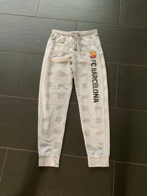 Gråa FC Barcelona mjukisbyxor - Snygga grå mjukisbyxor från FC Barcelona med tryckt klubbmärke och text längs ena benet. Byxorna har resår och dragsko i midjan samt muddar vid bensluten. Perfekta för chill eller träning, med all-over print av klubbens logga.