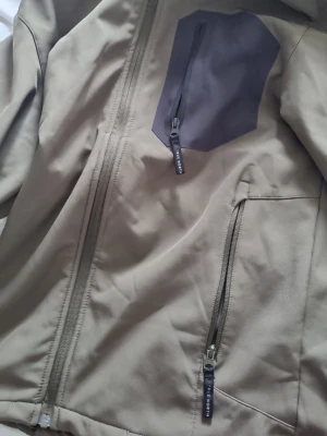 Beige vindjacka från Nordbjørn - Snygg beige vindjacka från Nordbjørn med dragkedja framtill, två fickor med dragkedja och en mörkgrå bröstficka. Jackan har en sportig look och är tillverkad i ett lätt material som skyddar mot vind.