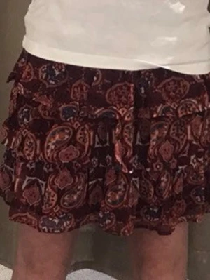 Mönstrad populär kortkjol i boho-stil - Säljer en kortkjol med bohemiskt paisleymönster i vinröda, orangea och blå toner. Väldigt populär kjol! Kjolen har volangdetaljer och är tillverkad i ett lätt, flödande tyg som ger en skön och avslappnad känsla. Perfekt för dig som gillar färg och mönster! Storlek 146/152 men passar mig som 172. Den är väldigt stretchig i midjan och blir inte för kort för mig. Endast andvänd få gånger och inga tecken på slitage