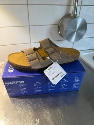 Birkenstock Arizona BS Taupe sandal - Säljer ett par Birkenstock Arizona BS sandaler i färgen taupe. De har två breda remmar med justerbara metallspännen och mjuk fotbädd. Ovandelen är i mocka och sulan är i kork och gummi för skön komfort. Perfekta för dig som gillar stilren och avslappnad look. Skorna är helt nya oanvända , i butik kostar dem 1900kr , säljs endast för 950kr . Fraktas samma dag . Först till kvarn storlek 43. 