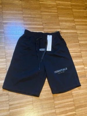 Svarta shorts från Essentials Fear of God - Svarta shorts från Essentials Fear of God med tryckt logga på vänster ben. Modellen har elastisk midja med snörning och är tillverkad i mjuk bomull. Perfekta för en avslappnad och trendig stil. Riktigt bra kvalité men den är lite för mig